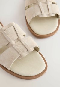 Beiga mockaskinnsandaler med två breda remmar, mjuk krämfärgad fotbädd och ljusbrun sula, med en minimalistisch design.