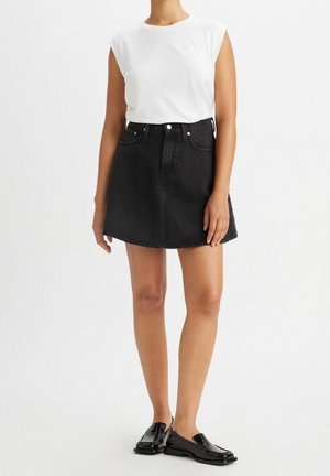 Femme portant un haut blanc sans manches, une jupe en jean noire taille haute et des mocassins en cuir noir, debout devant un fond uni.