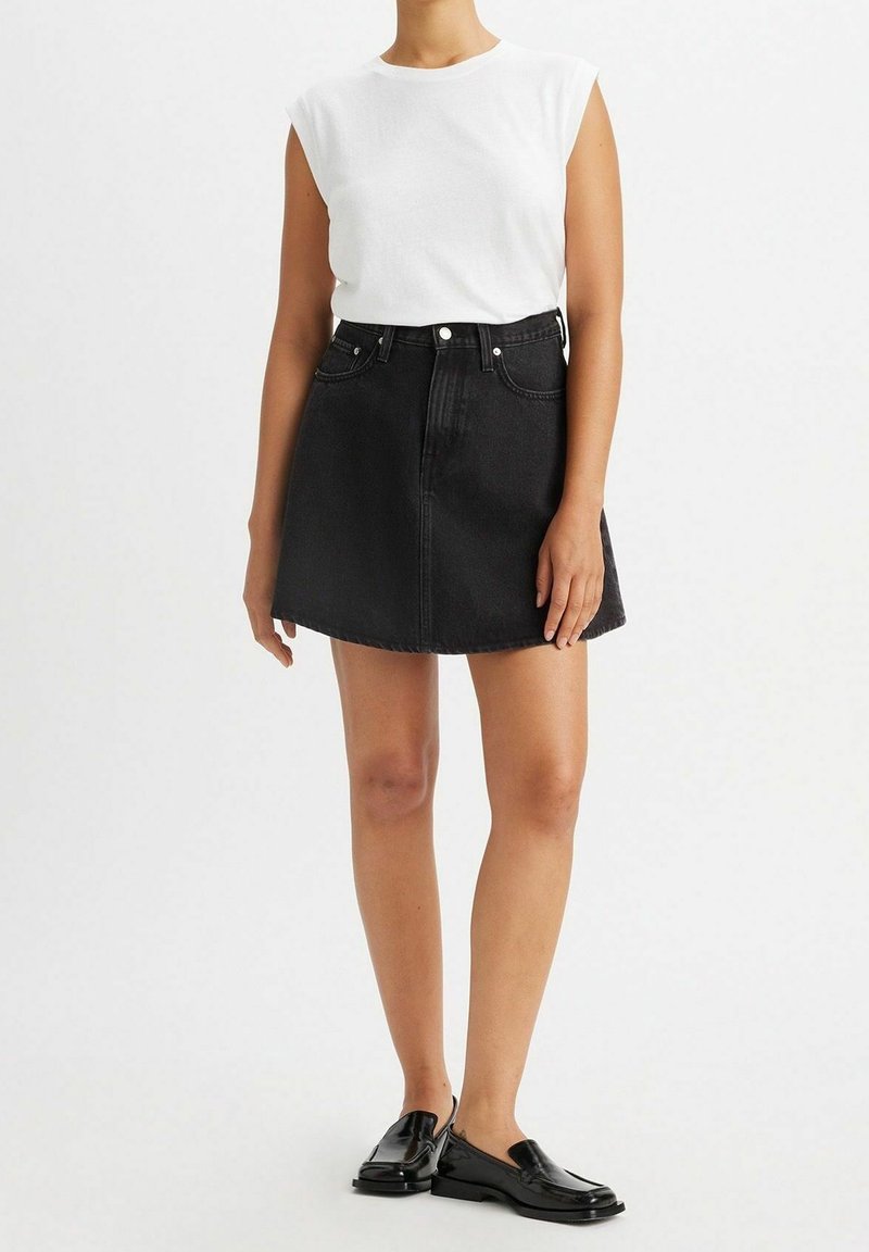 Femme portant un haut blanc sans manches, une jupe en jean noire taille haute et des mocassins en cuir noir, debout devant un fond uni.