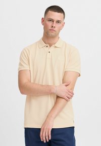 Beige polo skjorte med korte ermer og krave, med to brune knapper. Modellen har et sølvarmbånd og poserer foran en ensformig bakgrunn.