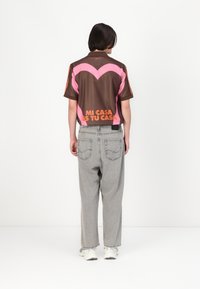 Person står vänd bort med en brun och rosa skjorta med texten "MI CASA ES TU CASA", grå jeans och vita sneakers mot en vit bakgrund.