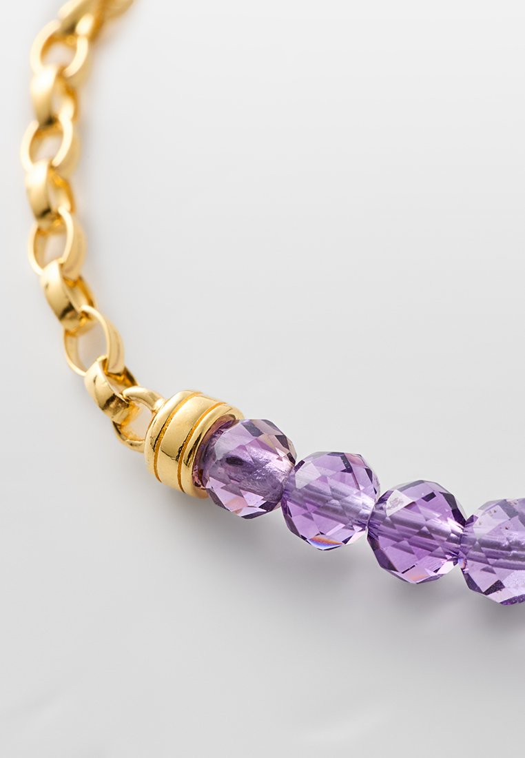 Segment eines goldenen Kettenarmbands mit facettierten runden lila Perlen, die neben einem goldenen Spacer mit zwei Ringen auf einem hellen Hintergrund aufgereiht sind.