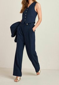 Vrouw in een marineblauwe mouwloze vest met knopen en bijpassende geplooide wijde broek, met gouden open teelt hakken, terwijl ze een marineblauwe jas vasthoudt.