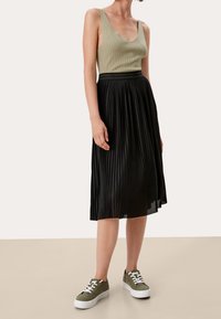 Gonna midi plissettata nera con vita liscia abbinata a un top senza maniche color oliva chiaro e sneakers verdi con suola e lacci bianchi.