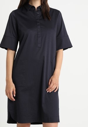Kobieta w granatowej sukience do kolan z krótkimi, szerokimi rękawami, kołnierzykiem i częściowym rozporkiem na guziki, stojąca na białym tle.