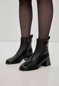 Cizme negre din piele până la glezne, cu un design elegant, fermoar pe lateral și toc pătrat. Accentuate cu o bareta cu cataramă metalică.