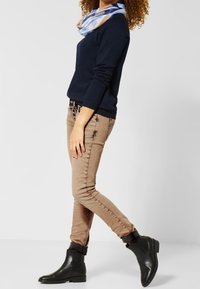 Un pull bleu marine et un jean skinny beige associés à des bottines noires. Une écharpe bleu clair à motifs est drapée autour du cou.