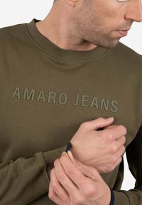Sudadera verde oliva con cuello y puños acanalados, con texto en relieve "AMARO JEANS" en color a juego. Se aprecia la textura suave de la tela.
