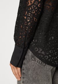 Black lace blouse with long sleeves, featuring a textured pattern and buttoned cuffs, worn over grey denim jeans showing pocket detail.