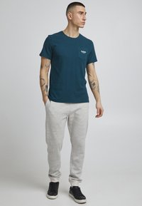 Camisa de manga corta en color azul petróleo con detalle de bolsillo, combinada con pantalones jogger gris claro. El modelo lleva zapatillas negras, mostrando un atuendo casual.
