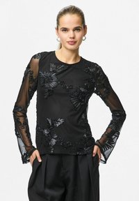 Kvinne iført svart gjennomsiktig langermet topp med blomsterapplikasjon og svarte bukser, hendene i lommene, håret trekt bakover og sølvfargede hoop-øreringer.