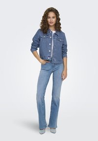 Veste en denim bleu moyen avec fermeture à boutons, deux poches poitrine et manches longues. Associée à un jean évasé bleu clair et des chaussures grises.