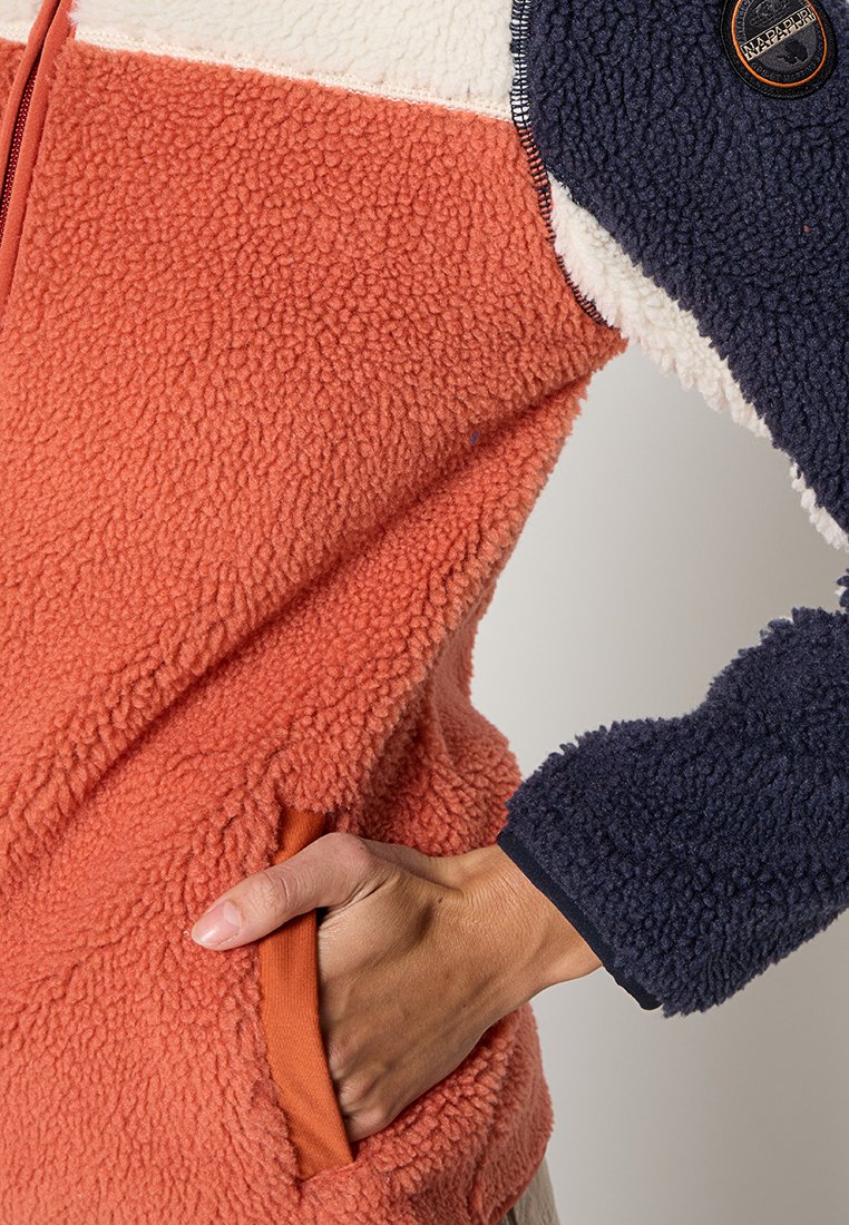 Chaqueta de forro polar con paneles en naranja, crema y azul marino. Tejido texturizado con cremallera y bolsillo lateral; parche de logo visible en la manga azul marino.
