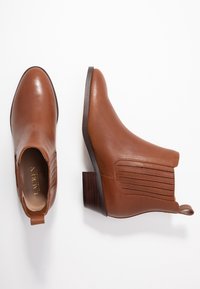 Botines de cuero marrón con textura suave, punta redondeada, paneles laterales elásticos y tacón apilado. Cuentan con una lengüeta para facilitar su uso.