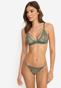 Σετ σουτιέν με δαντέλα και στρινγκ σε σκούρο πράσινο χρώμα. Διαθέτει τριγωνικά κύπελλα με λουλουδάτο μοτίβο, λεπτές τιράντες και λεπτή, διαφανή υφή.