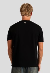 Czarna bawełniana koszulka z krótkim rękawem widziana z tyłu, z okrągłym dekoltem. Na karku znajduje się metalowy akcent z logo.