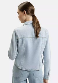 Jasnoniebieska dżinsowa kurtka o dopasowanym kroju, z przednimi kieszeniami i zapięciem na guziki. Tkanina ma lekko widoczną teksturę, z przeszytymi szwami.
