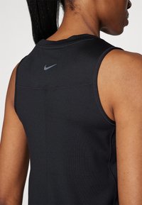 Donna con pelle scura e coda di cavallo che indossa una canotta nera senza maniche con il logo grigio della Nike sulla parte superiore della schiena.