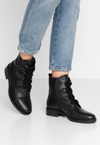 Bottines en cuir noir avec des lacets épais, des accents métalliques et une fermeture éclair sur le côté, dotées d'un talon bas texturé et d'une semelle en caoutchouc.