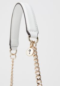 Hvid tekstureret faux læderrem med guldfarvet hardware, der har en kæde med en rund charm mærket med et spørgsmålstegn.