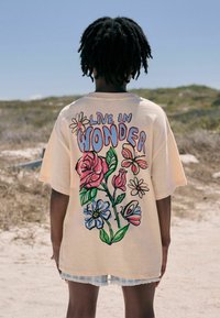 Kortærmet, oversized beige t-shirt med et flerfarvet blomsterdesign og sætningen "LEV I UNDERDEN" tydeligt på ryggen.