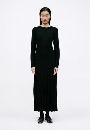 Fekete bordázott hosszú ujjú ruha kör alakú nyakkivágással és testhez simuló derékkal, amely a bokákig ér. A modellen fekete harisnya és klumpák vannak.