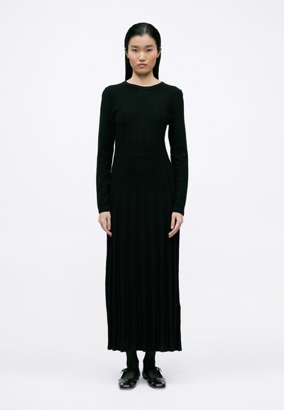 Schwarzes, geripptes Langarmkleid mit rundem Ausschnitt und taillierter Form, das bis zu den Knöcheln reicht. Das Model trägt schwarze Strumpfhosen und Clogs.
