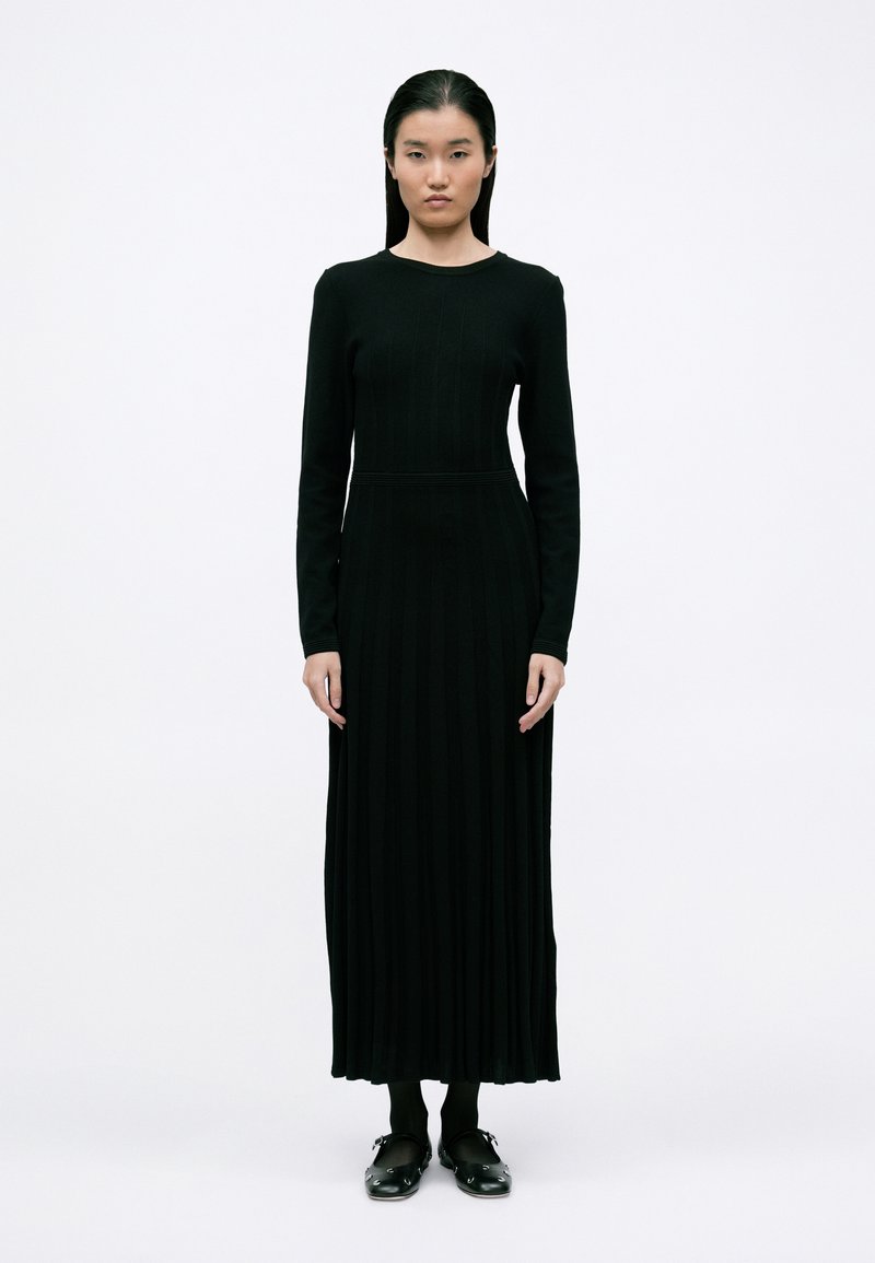 Vestido preto de mangas longas com textura em ribas, de decote redondo e cintura ajustada, que se estende até os tornozelos. A modelo usa meias pretas e tamancos.