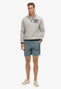 Grijze rits-sweatshirt met blauwe tekst, gecombineerd met turquoise shorts met een logo-opdruk. Model draagt lichtgekleurde sneakers.