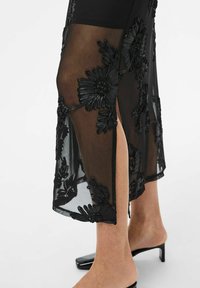 Robe en dentelle noire avec des détails de broderie florale, tissu transparent, fente haute et silhouette ajustée. Mules noires à talons hauts visibles.