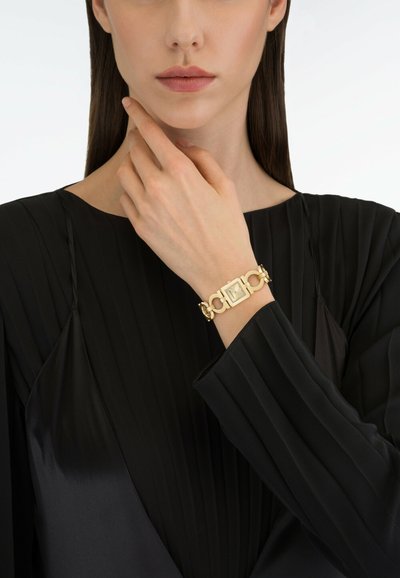 Frau trägt eine goldene Armbanduhr mit Gliederarmband am Handgelenk, bekleidet mit einem schwarzen, langärmeligen, plissierten Oberteil, Hand nahe am Kinn.