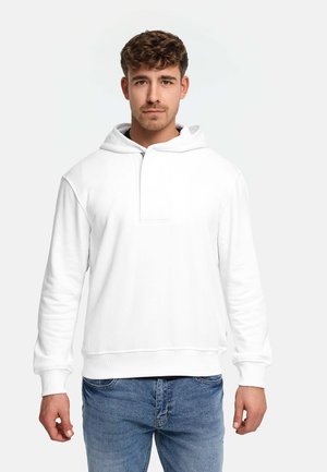 Mann mit kurzem braunen Haar, der einen weißen Kapuzenpullover und blaue Jeans trägt, steht vor einem schlichten hellen Hintergrund.