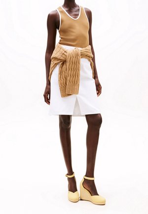 Haut sans manches beige associé à une jupe blanche avec une fente frontale, et un pull texturé beige noué autour de la taille ; chaussures à talons compensés jaunes.