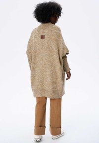 Długi, oversize sweter w beżowym i kremowym kole, z prążkowanymi mankietami i wysokim kołnierzem. Zawiera małą skórzaną naszywkę z logo z tyłu.