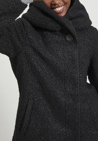 Svart texturerad kappa med en stor krage och en enda knappstängning, med sidofickor och synliga sömmar.