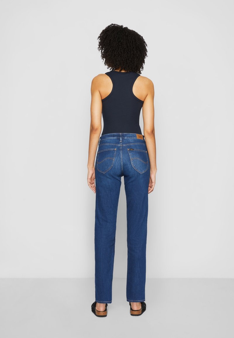 Jean en denim bleu foncé à coupe droite, avec poches arrière et étiquette de marque marron à la ceinture, porté avec un débardeur noir.