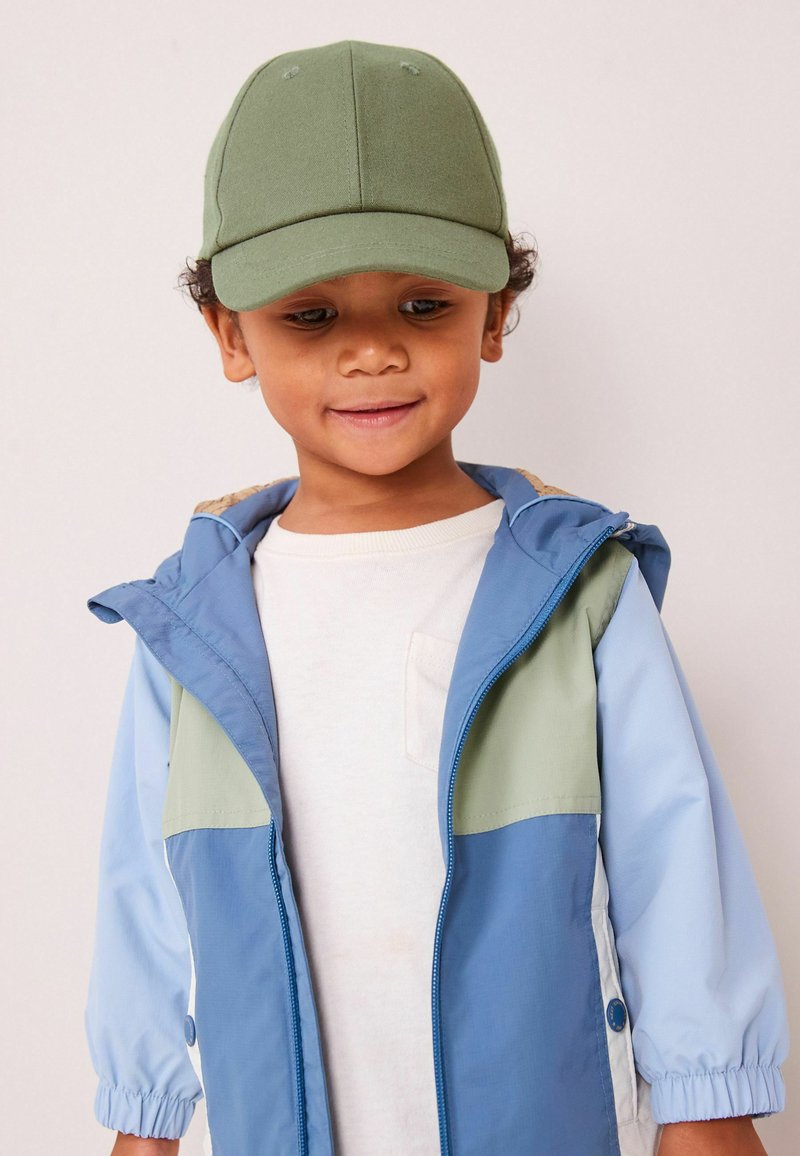 Cappello verde, giacca azzurro chiaro e verde oliva con polsini elasticizzati, sopra una maglietta bianca. Presenta una piccola tasca sulla maglietta e dettagli con zip.