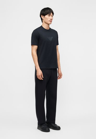 Νεαρός άνδρας στέκεται φορώντας μαύρο μπλουζάκι με διακριτικό λογότυπο, μαύρο παντελόνι και μαύρα αθλητικά παπούτσια, σε απλό ανοιχτό φόντο.