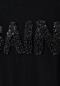 Camiseta de tela negra con adornos de cuentas y cristales brillantes en relieve formando la palabra "SAINT". Textura suave.