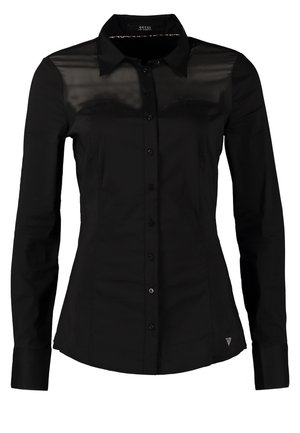Femme portant un chemisier noir boutonné avec un détail transparent en haut, un pantalon beige slim et des bottines noires, tenant une pochette noire dans sa main gauche.
