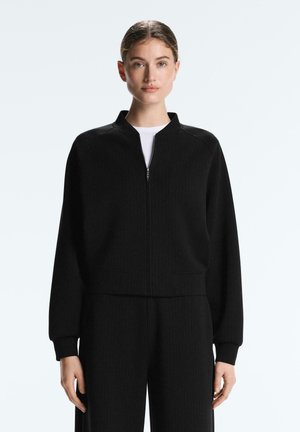 Donna in piedi, che indossa una giacca nera con zip e pantaloni abbinati sopra una maglietta bianca, su uno sfondo chiaro e uniforme.