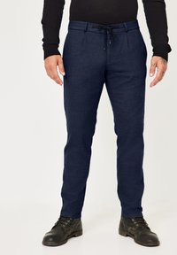 Marineblaue, maßgeschneiderte Hosen mit einem Kordelzugbund, vorderen Falten und glatter Textur. Kombiniert mit schwarzen, geschnürten Stiefeln.