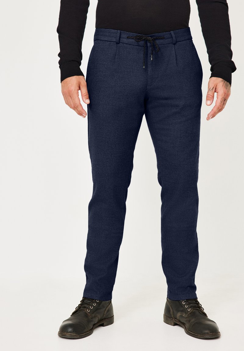 Marineblaue, maßgeschneiderte Hosen mit einem Kordelzugbund, vorderen Falten und glatter Textur. Kombiniert mit schwarzen, geschnürten Stiefeln.