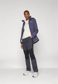 Γυναίκα με ναυτική αδιάβροχη ζακέτα, λευκό μπλουζάκι, σκούρους παντελόνες με μαύρες επιφάνειες και λευκά αθλητικά παπούτσια, που στέκεται σε απλό φόντο.