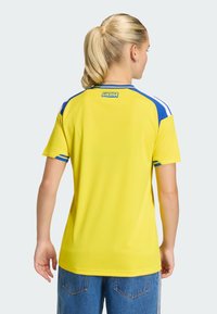Gelbes Sporttrikot mit blauen Akzenten; verfügt über Streifen auf den Schultern, kurze Ärmel und den Schriftzug „SVERIGE“ auf dem oberen Rücken. Glatte Stofftextur.