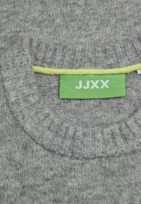 Pilka megztinė su iškilniu apykakle, turinti ryškiai geltoną pamušalą šalia ženklo, kuriame baltu raštu užrašyta "JJXX" ant žalio fono.