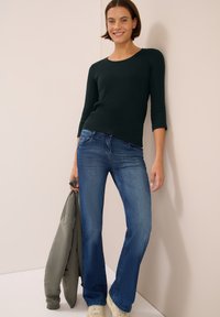 Dunkelgrünes, langärmliges Oberteil kombiniert mit blauen, ausgestellten Jeans. Das Model hält eine graue Jacke und trägt cremefarbene Sneaker. Einfaches, lässiges Outfit.