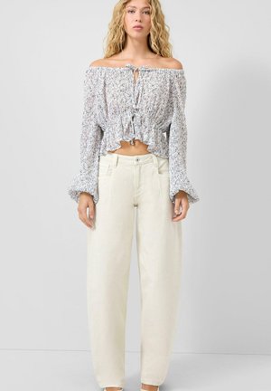 Vrouw die een witte, bloemenoff-shoulder blouse met lange mouwen en voorste strikjes draagt, gecombineerd met losse, crèmekleurige, hooggetailleerde broek met wijde pijpen, staande stil.