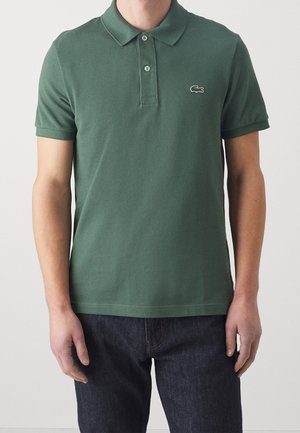 Mann trägt ein dunkelgrünes kurzärmeliges Polohemd mit zwei Knöpfen und einem kleinen gestickten Logo auf der linken Brust.