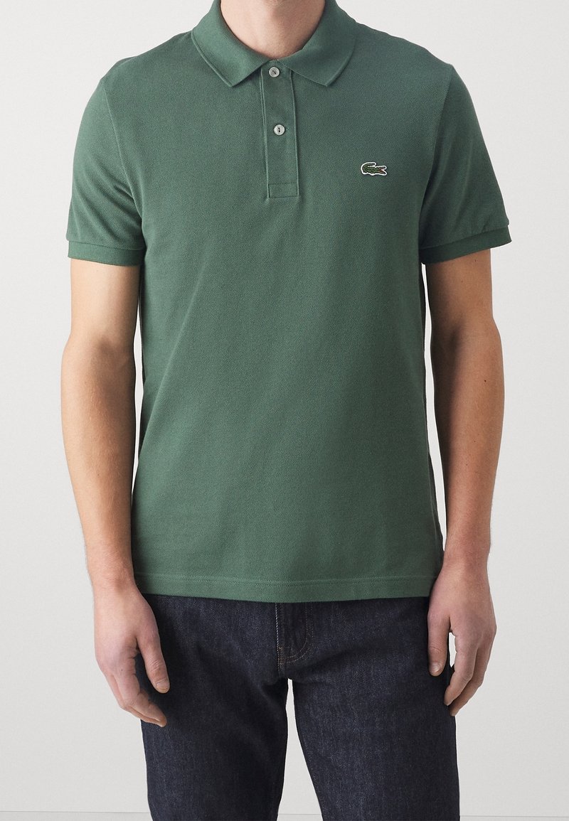 Homme portant un polo vert foncé à manches courtes avec deux boutons et un petit logo brodé sur la poitrine gauche.